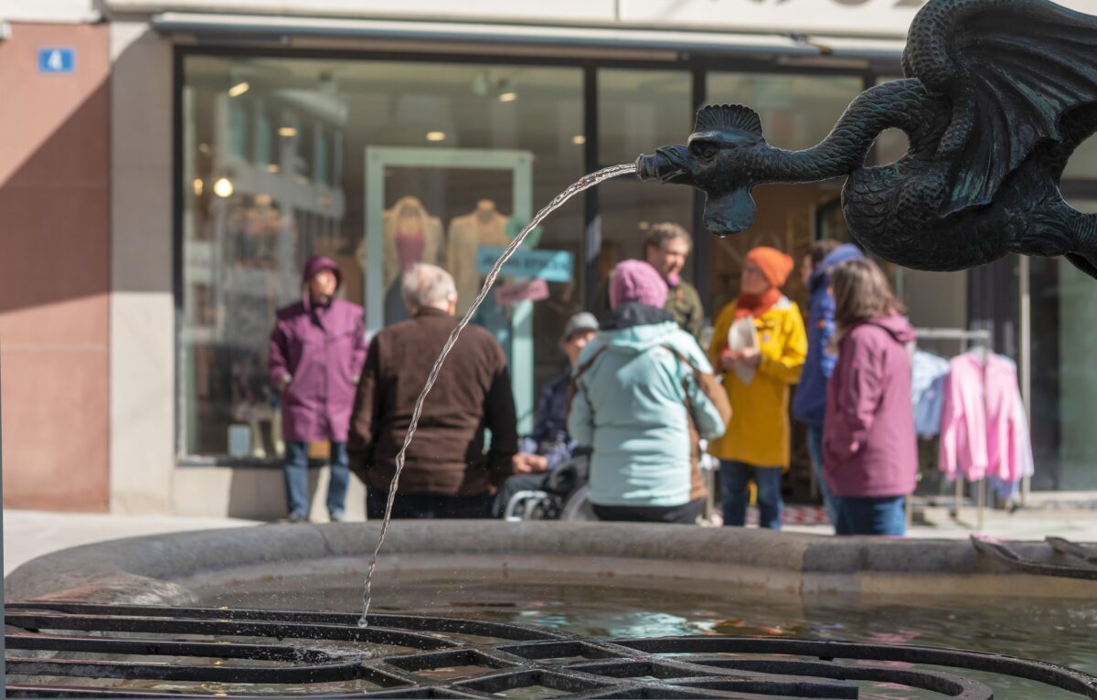 Ein Drache mit geöffnetem Maul, aus dem Wasser in einen antiken Brunnen fliesst. Im Hintergrund unscharf zu erkennen eine Gruppe von 8 Personen, gekleidet in farbigen Jacken und Mützen.