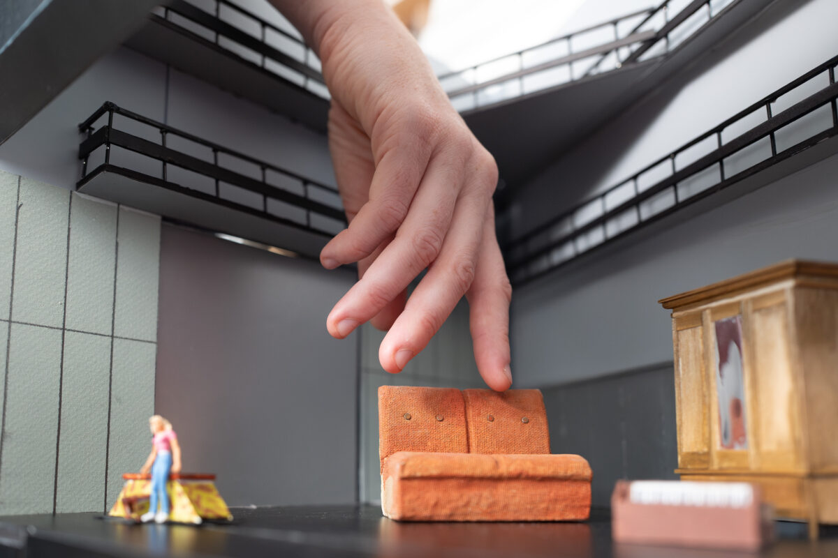 Es ist eine verkleinerte Bühne mit einem Bühnenbild zu sehen. Das ist ein Modell. Eine Hand geht in das Modell. Die Hand tastet durch den Bühnenraum. Die Hand ertastet ein Sofa, ein Klavier und die Bühne.