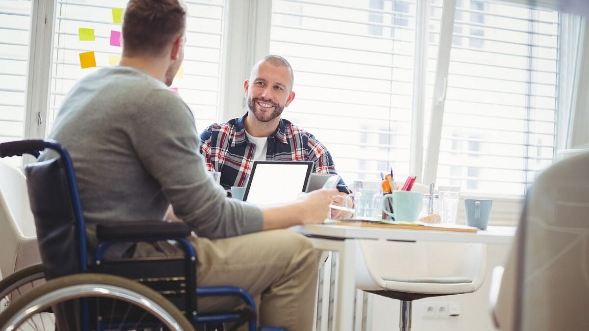 Mitarbeitender im Rollstuhl diskutiert mit einem Arbeitskollegen an einem Besprechungstisch. Auf dem Tisch aufgeklappte Laptops, Schreibutensilien, Tassen und Gläser.