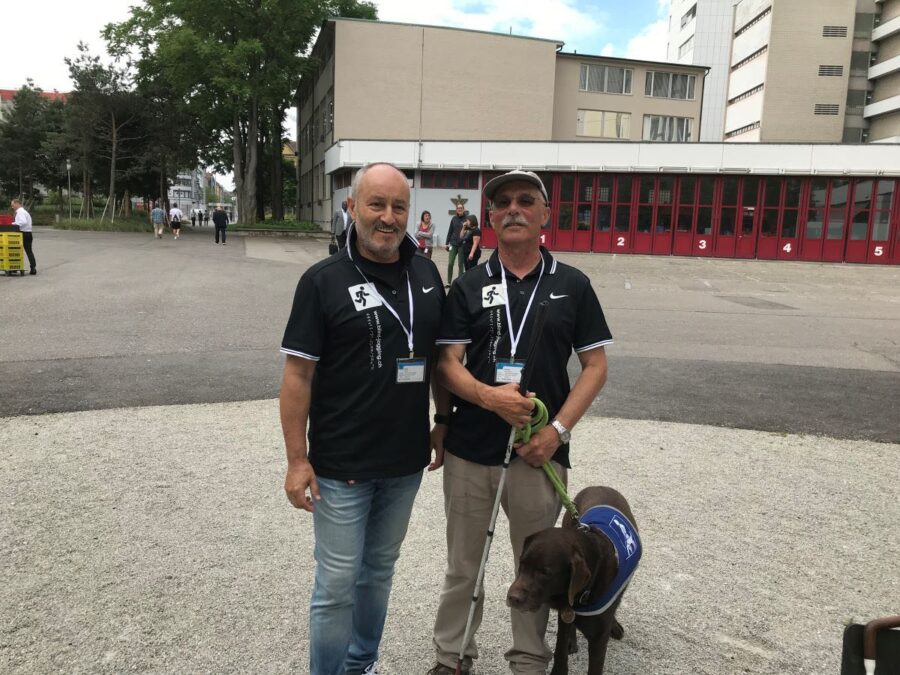 Wir sehen einen blinden Mann mit Begleithund und einen Begleiter.