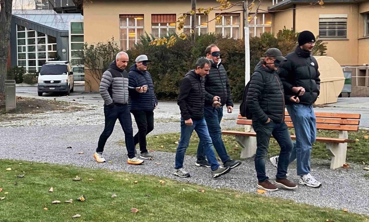 Guides führen sehende Menschen mit der Dunkelbrille auf einen Weg.
