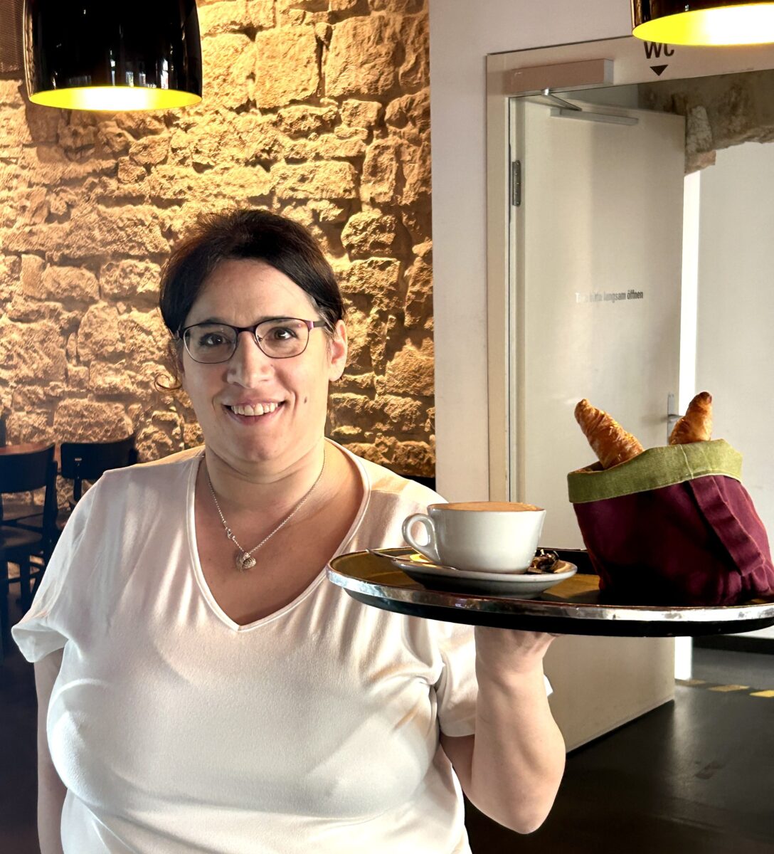 Eine Frau hält ein Servier-Tablet in der Hand. Auf dem Servier-Tablet hat es eine Tasse Kaffee und ein Beutel mit zwei Gipfeli.