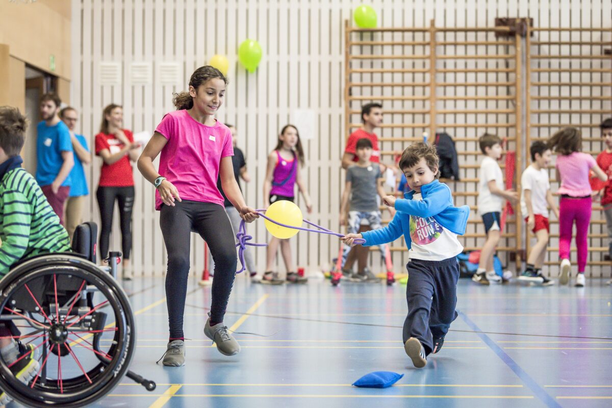 Kinder mit und ohne Behinderung absolvieren gemeinsam Stafetten in einer Turnhalle. Dabei sind sie durch ein Seil verbunden, mit dem sie einen Ballon transportieren müssen. Die anderen Kinder im Hintergrund warten gespannt auf ihren Einsatz.