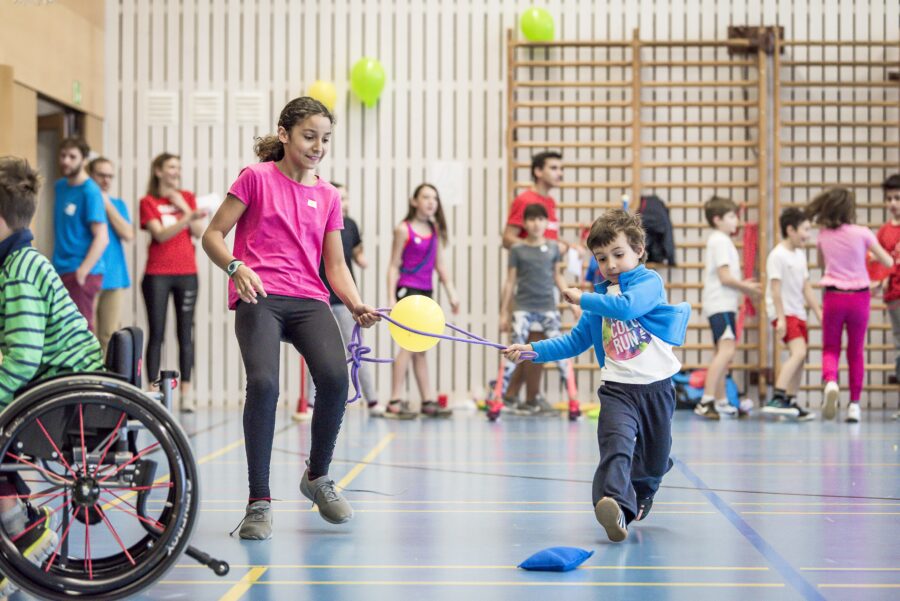 Kinder mit und ohne Behinderung absolvieren gemeinsam Stafetten in einer Turnhalle. Dabei sind sie durch ein Seil verbunden, mit dem sie einen Ballon transportieren müssen. Die anderen Kinder im Hintergrund warten gespannt auf ihren Einsatz.