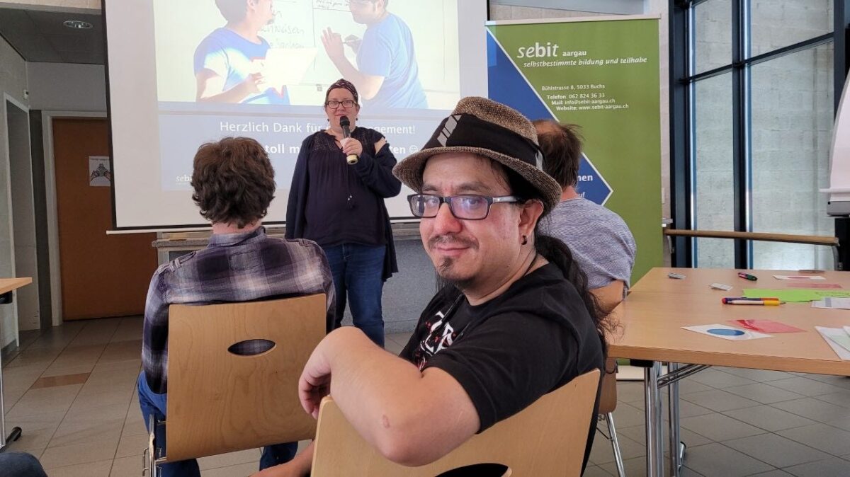 Zu sehen ist ein sitzender Mann. Er trägt eine Brille und einen Hut. Er blickt in die Kamera. Dahinter sind zwei Personen zu sehen, die in die entgegengestzte Richtung blicken, in Richtung Leinwand. Vor der Leinwand steht eine Frau mit Mikrophon. Sie moderiert einen Workshop.