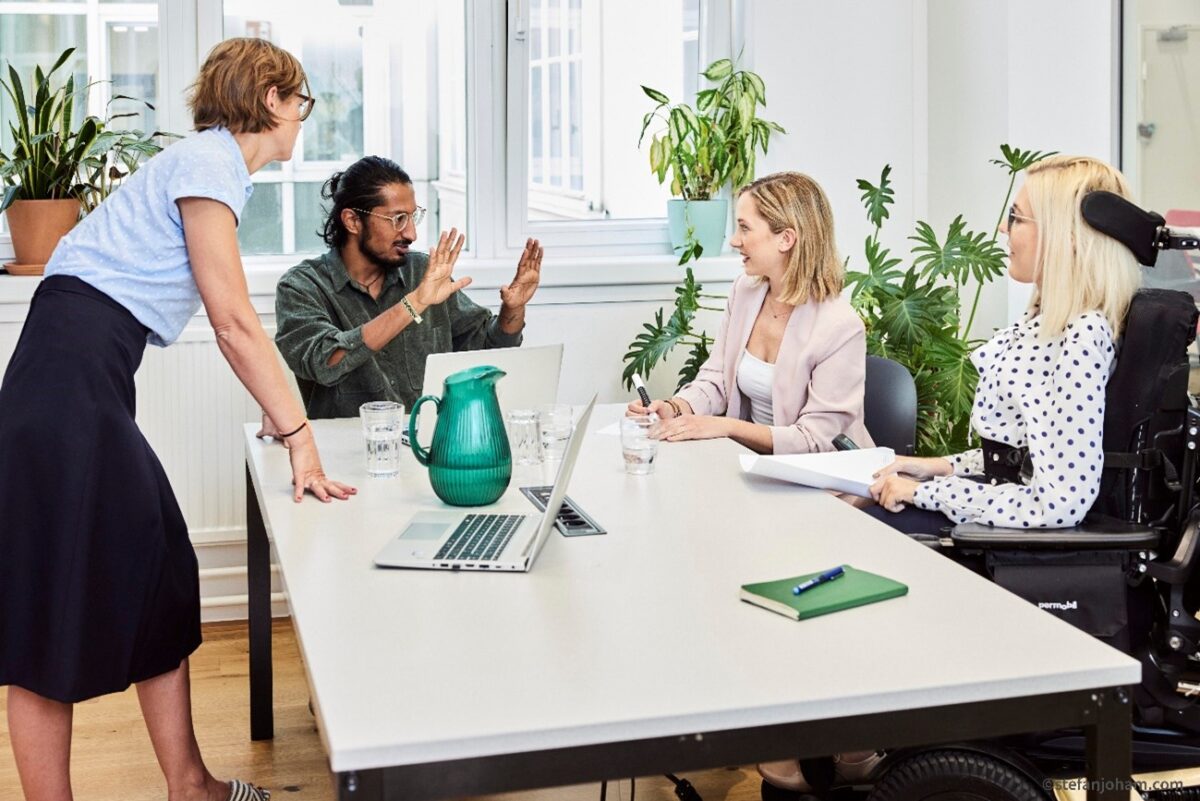Eine Arbeitssituation in einem Büro. Drei Damen und ein Herr sprechen miteinander. Die Dame links steht neben dem Tisch, der Herr daneben sowie zwei weitere Damen sitzen. Der Herr kommuniziert in Gebärdensprache. Die Dame ganz rechts ist nutzt einen Elektrorollstuhl. Auf dem Tisch befinden sich unter anderem ein Laptop und ein Notizbuch.