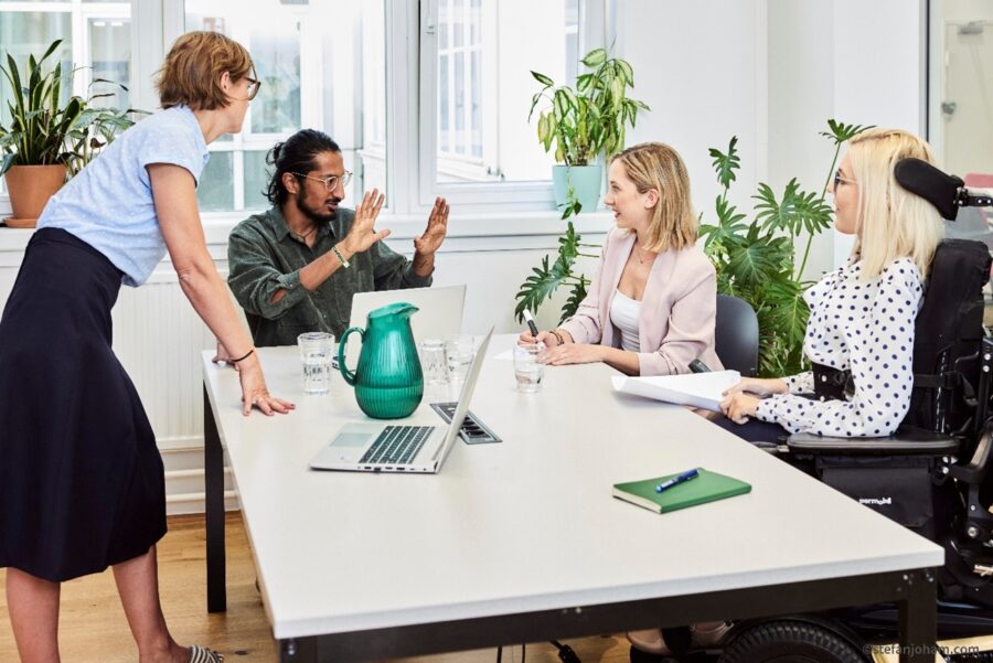 Eine Arbeitssituation in einem Büro. Drei Damen und ein Herr sprechen miteinander. Die Dame links steht neben dem Tisch, der Herr daneben sowie zwei weitere Damen sitzen. Der Herr kommuniziert in Gebärdensprache. Die Dame ganz rechts ist nutzt einen Elektrorollstuhl. Auf dem Tisch befinden sich unter anderem ein Laptop und ein Notizbuch.