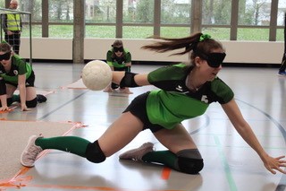 Eine Torballspielerin mit dem TB Glarus 11-Trikot wirft den Ball aus dem Sitzen auf die andere Feldseite. Die Spielerin trägt eine Dunkelbrille und Schutzkleidung (Knie- und Ellbogenschoner). Im Hintergrund sind die beiden weiteren Spielerinnen des Teams zu sehen.