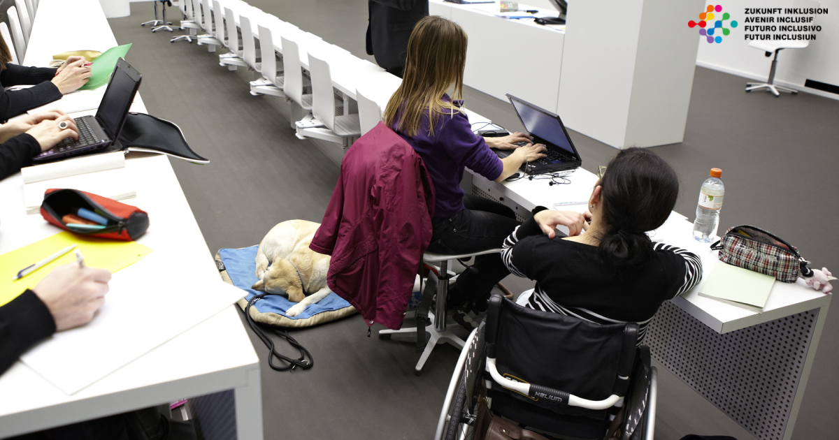 Zwei Frauen sitzen in einem Hörsaal. Eine Frau hat einen Begleithund. Die andere Frau sitzt im Rollstuhl.