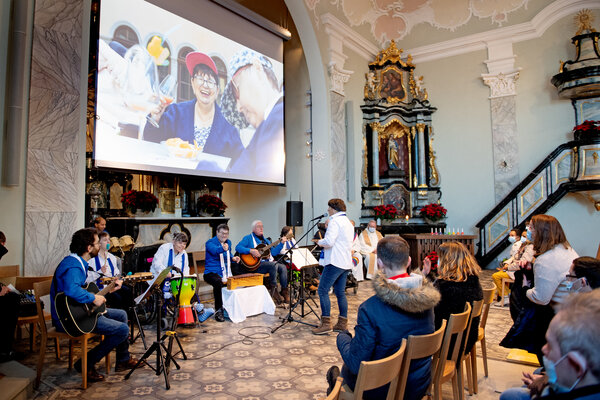Auf dem Bild ist der Innenraum der Klosterkirche Rathausen zu sehen. Die «SSBL-Musig Bänd» musiziert. Zuschauer hören zu. Vor dem Hauptaltar ist eine grosse Leinwand, auf der ein Bild von einer fröhlichen Frau projiziert ist.