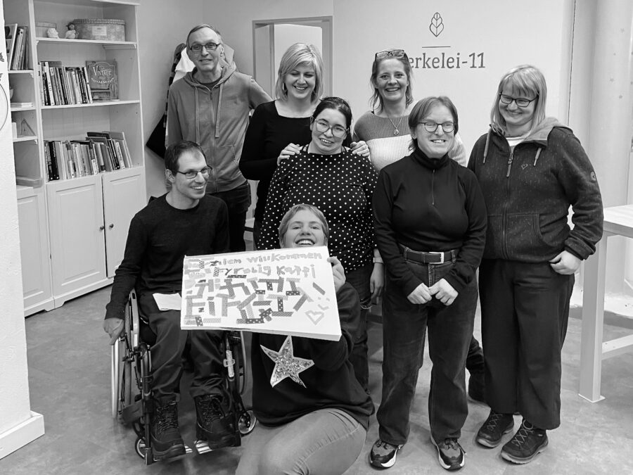 Eine Gruppe von acht Menschen haben sich in einem Innenraum in drei Reihen platziert, die vorderste Person zeigt ein selbst gestaltetes Plakat, darauf steht: «Herzlich Willkommen zum Fyrobig Kaffi!» Die Menschen lachen uns freundlich an. Hinten an der Wand ist ein Schriftzug: Werkelei-11, das ist der Name des Ortes.