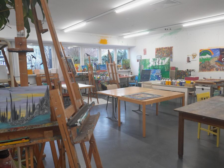 Diverse Arbeitstisch und Staffeleien umgeben von vielen kunstvollen und farbenprächtigen Bildern im offenen Atelier
