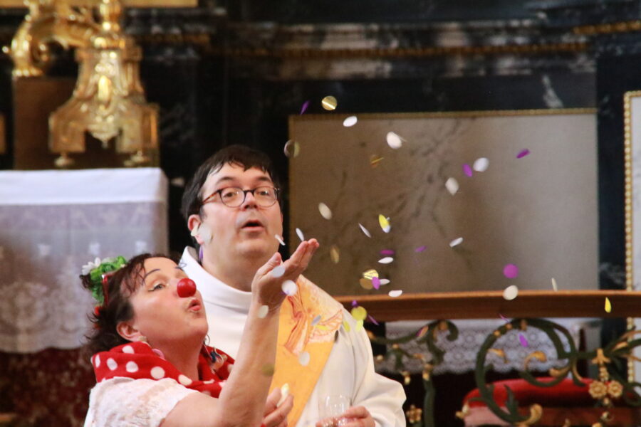 Es sind die Gesichter von zwei Menschen in einer Kirche zu sehen. Ein Gesicht einer Clownin und ein Gesicht eines Seelsorgers/Diakons. Aus der Hand der Clownin fliegen kleine bunte runde Schnipsel in die Luft. Die beiden staunen.
