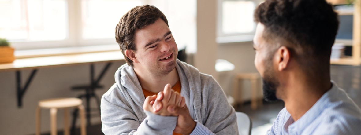 Zwei lachende junge Männer im Handschlag, sitzen sich gegenüber.