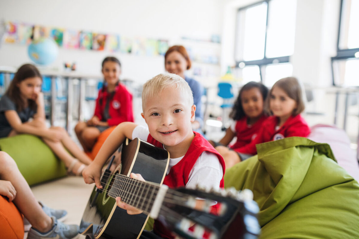 Ein lachendes Kind, was eine Gitarre in der Hand hält. Das Kind sitzt in einer Gruppe von vielen Kindern und einen Erwachsenen.