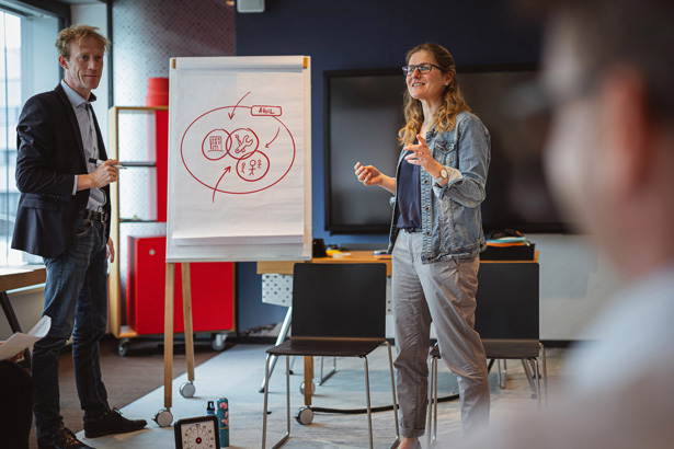Zwei Personen diskutieren vor einem Flipchart im modernen Klassenraum. Verschwommen sieht man weitere Personen, die der Diskussion zuhören.