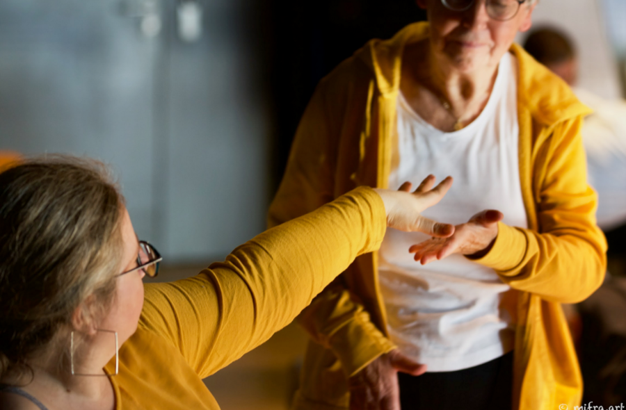 Im Vordergrund links sitzt eine Frau, die ihre Hand nach hinten zu einer stehenden Frau ausstreckt. Die stehende Frau nähert sich mit ihrer Hand der vorderen Frau an. Sie nehmen miteinander Kontakt auf. ©mifra.art