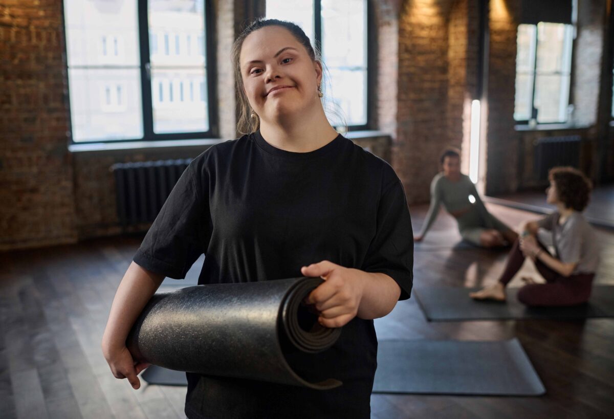 Eine junge Frau mit Down Syndrom hält eine schwarze Yoga Matte. Die Frau trägt dunkle Sportkleider und hat lange dunkle Haare. Sie befindet sich in einem Kursraum zusammen mit anderen Personen. Diese sitzen im Hintergrund und reden.
