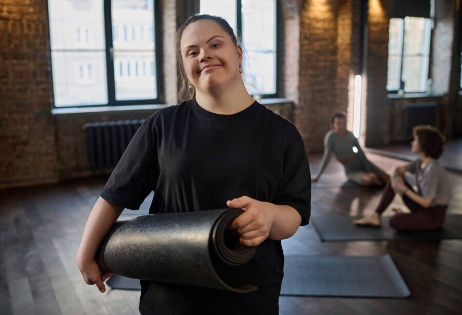 Eine junge Frau mit Down Syndrom hält eine schwarze Yoga Matte. Die Frau trägt dunkle Sportkleider und hat lange dunkle Haare. Sie befindet sich in einem Kursraum zusammen mit anderen Personen. Diese sitzen im Hintergrund und reden.