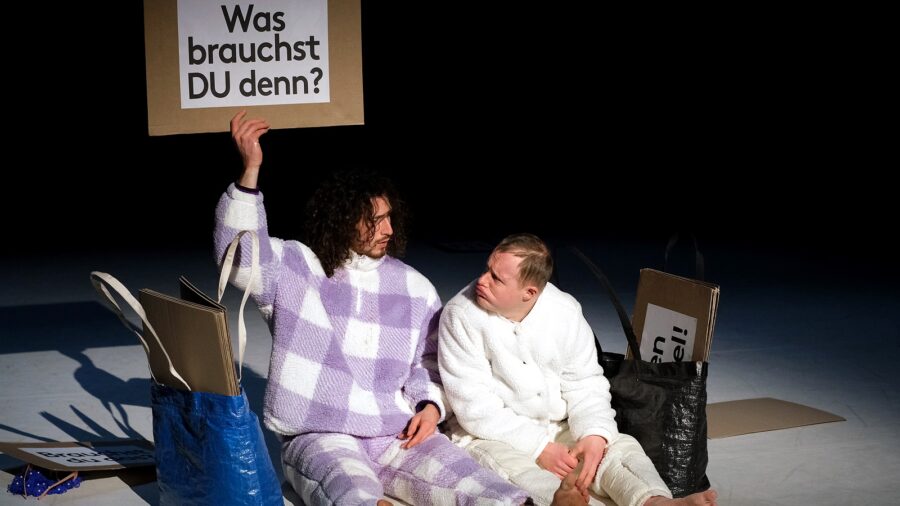 Das Bild zeigt zwei männlich gelesene Personen nebeneinander sitzend auf dem Boden. Sie schauen einander an. Der Hintergrund ist schwarz, es werden nur die beiden Personen mit einem Spot ausgeleuchtet. Die linke Person trägt einen weiß, lila quadratisch gemusterten Zweiteiler und hält ein Pappschild nach oben. Auf dem Schild steht „Was brauchst DU denn?“. Die Person rechts trägt einen weißen Zweiteiler und schaut leicht launisch zu der anderen Person. Neben beiden Personen steht je eine Plastiktasche mit mehreren Pappschildern. Wenige Pappschilder mit Aufschriften liegen auf dem Boden um die Personen herum. Die Aufschriften sind nicht lesbar.