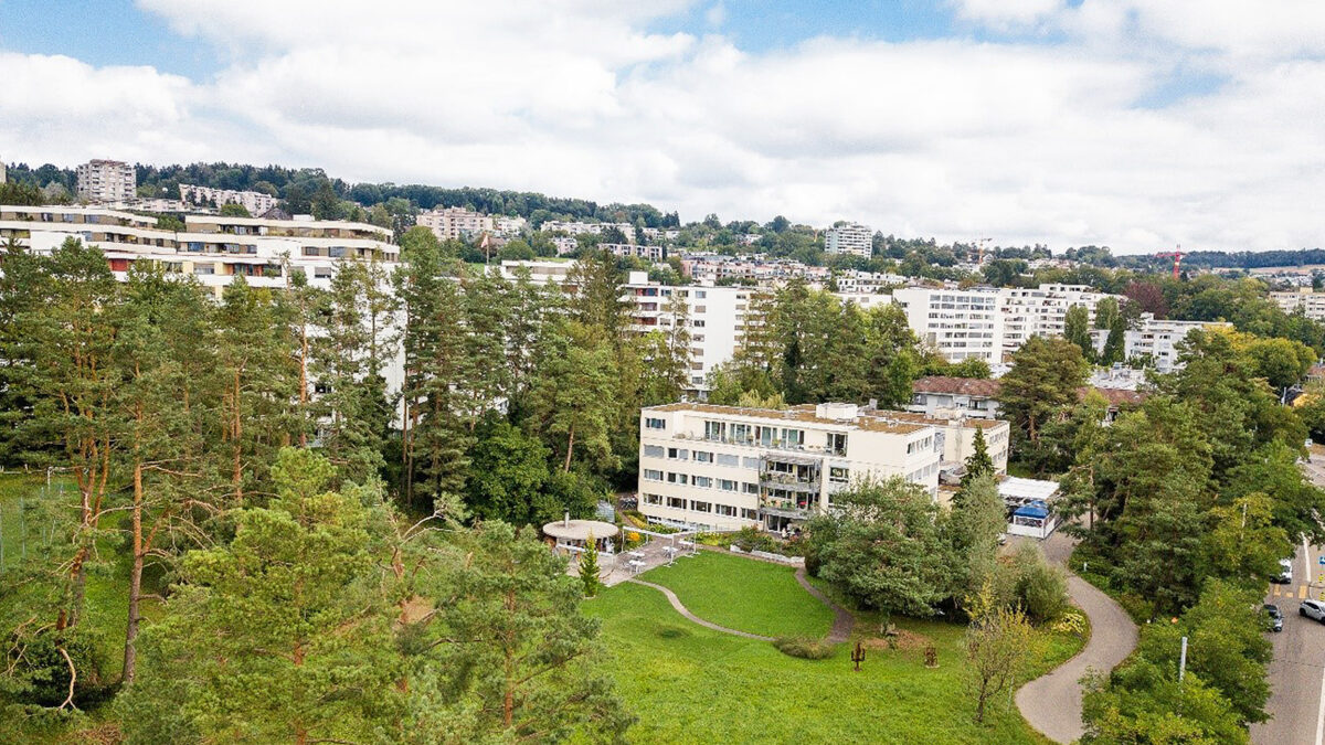 Auf dem Bild ist der Garten des Wohnzentrum Frankentals in hellem Frühlingssonnenlicht zu sehen aus der Vogelperspektive zu sehen. Der Garten erscheint in frischem Grün und lädt zum Spazieren und Verweilen ein. Ein Weg durch die idyllische Landschaft ist sichtbar. An diversen Stellen sind Sitzmöglichkeiten wie Bänke und hinter einer Baumgruppe befindet sich ein Teich. In der Mitte des Bildes ist das Wohnzentrum Frankental zu sehen und im Hintergrund ist der Hönggerberg erkennbar.