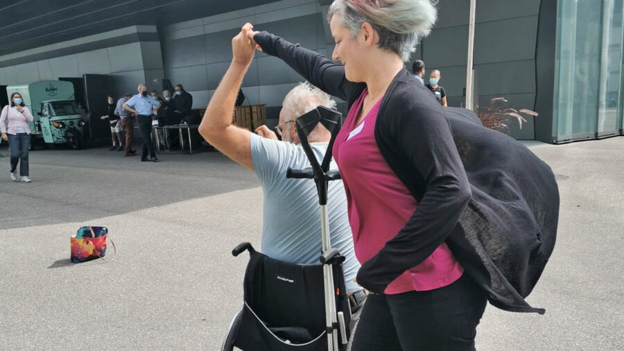 Ein Mann, der im Rollstuhl sitzt, hält die Hand einer Frau. Im Hintergrund ist ein Gebäude zu sehen.