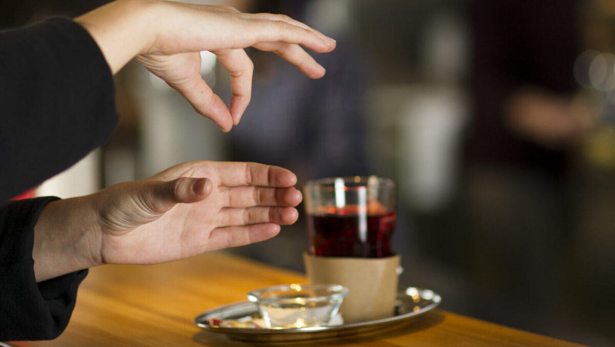 Gebärde für das Wort: «Tee»: Auf einem Tresen steht eine Teetasse. Daneben sind zwei Hände zu sehen. Die untere Hand zeigt geschlossene, gebeugte Finger mit abgespreiztem Daumen, während die andere Hand mit sich berührenden Daumen- und Zeigefingerspitzen darüber schwebt.