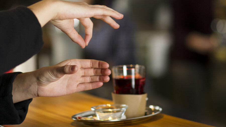 Gebärde für das Wort: «Tee»: Auf einem Tresen steht eine Teetasse. Daneben sind zwei Hände zu sehen. Die untere Hand zeigt geschlossene, gebeugte Finger mit abgespreiztem Daumen, während die andere Hand mit sich berührenden Daumen- und Zeigefingerspitzen darüber schwebt.