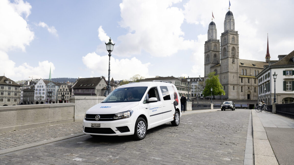 Ein weisses Auto mit der Aufschrift "Tixi Zürich". Dieses fährt durch die Stadt Zürich. Im Hintergrund ist das Grossmünster.