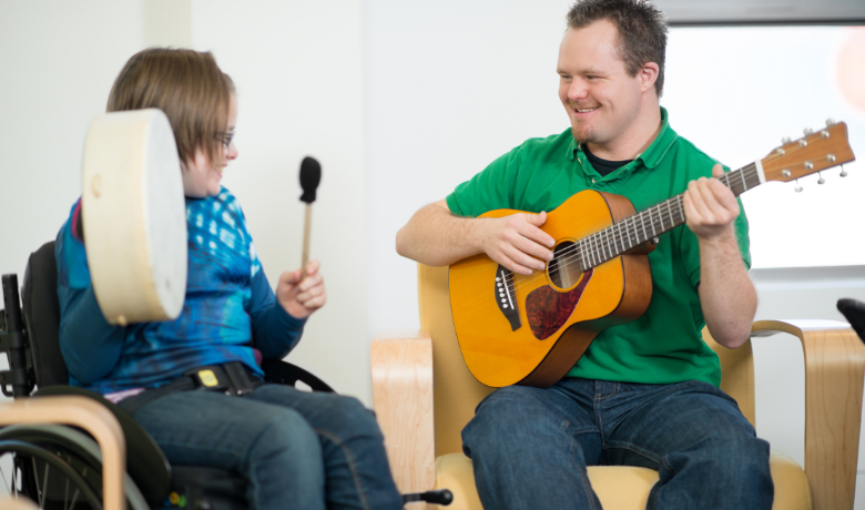 Gemeinsam musizieren: Mädchen im Rollstuhl spielt Tambourin, ein junger Mann mit Trisomie 21 spielt Gitarre.