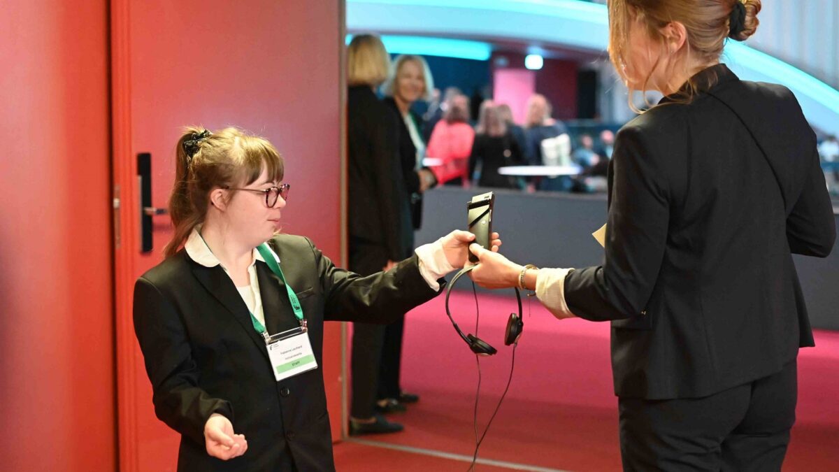 Eine junge Frau mit Downsyndrom reicht einer Kongressbesucherin die Kopfhörer mit Empfänger für die Simultanübersetzung. Beide Frauen tragen eine weisse Bluse und einen schwarzen Blazer.