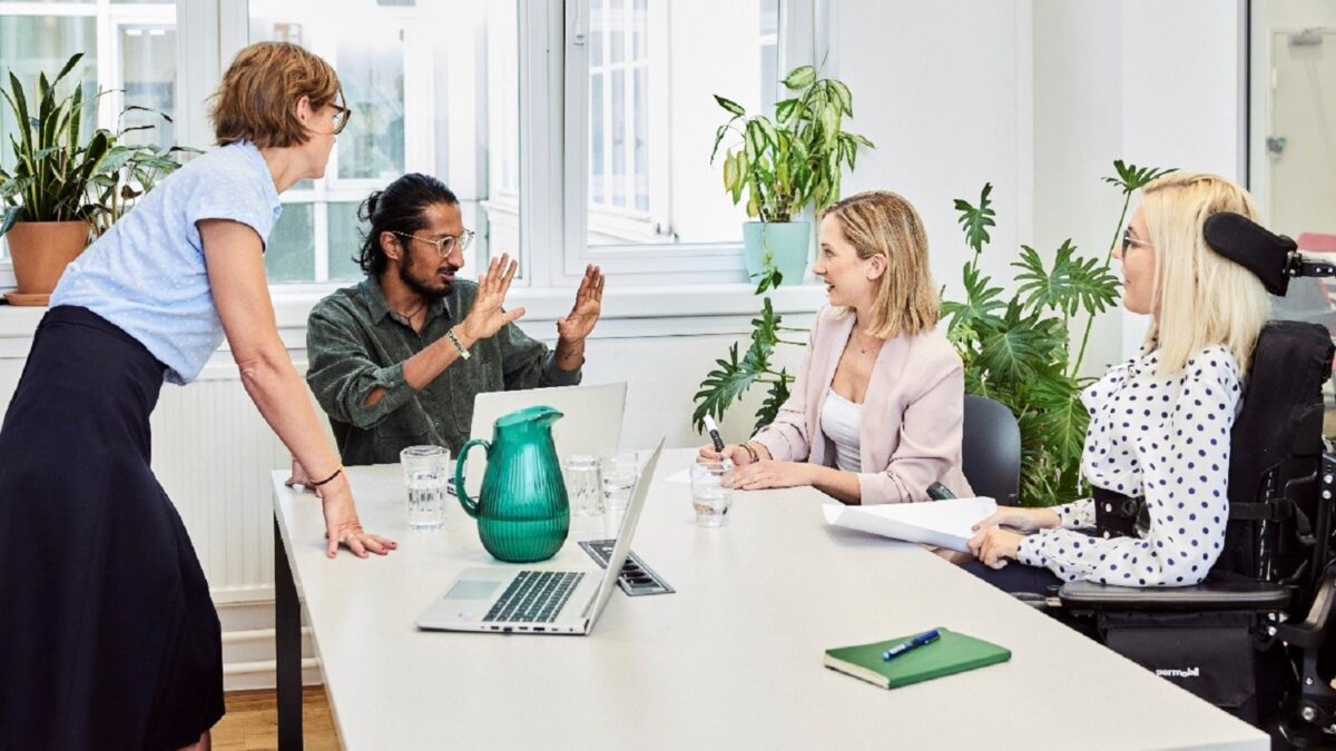 Eine Arbeitssituation in einem Büro. Drei Damen und ein Herr sprechen miteinander. Die Dame links steht neben dem Tisch, der Herr daneben sowie zwei weitere Damen sitzen. Der Herr kommuniziert in Gebärdensprache. Die Dame ganz rechts ist nutzt einen Elektrorollstuhl. Auf dem Tisch befinden sich unter anderem ein Laptop und ein Notizbuch.
