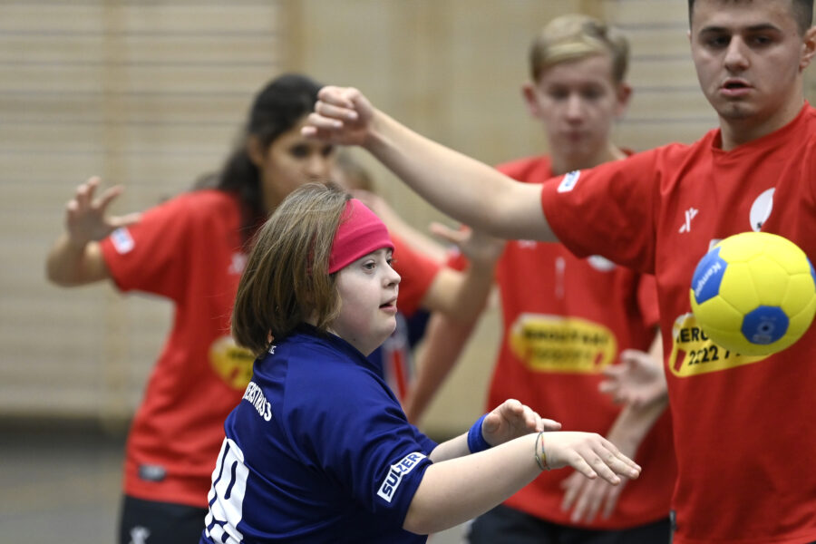 Zwei Teams spielen in der inklusiven Handball-Liga ein Spiel. SV Fides St. Gallen spielen in roten Trikots. Eine Athletin mit schwarzen Haaren und zwei Athleten sind abgebildet und verteidigen. Ein Athletin vom TV Unterstrass in blauem Trikots wirft gerade einen gelb-blauen Ball.