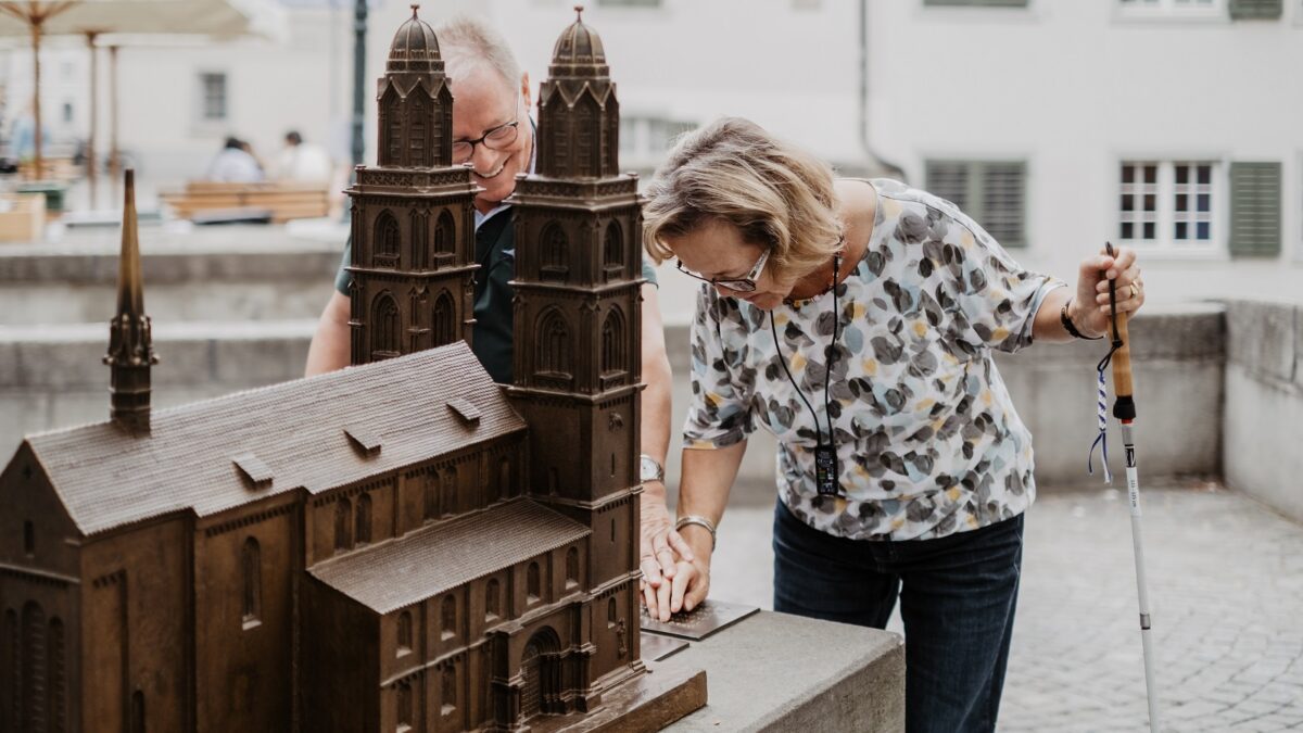Ein Mann und eine Frau mit einer Sehbehinderung ertasten das taktile Modell des Grossmünsters und lesen den Beschrieb in Braille.