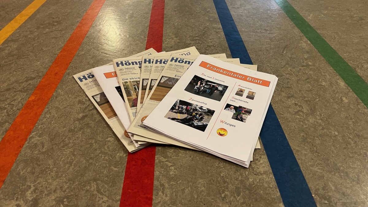 Es sind die Es sind sieben Zeitungen zu sehen, aufgefächert wie Jasskarten. gemischt 5x Höngger Zeitung und 2x Frankentaler Blatt. Sie liegen auf einem Boden mit eingelassenen senkrechten farbigen Linien, die nach hinten parallel zusammenlaufen, ähnlich wie bei einer Kegel- oder Bowlingbahn.