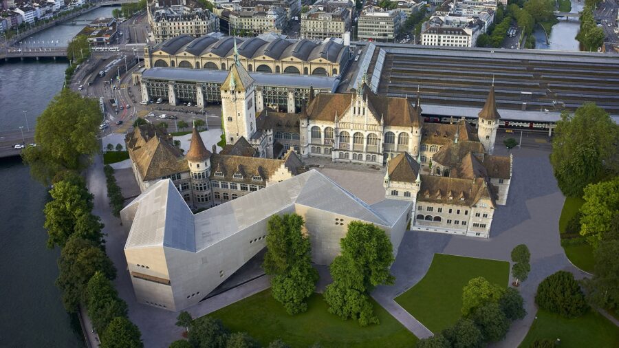 Landesmuseum und Hauptbahnhof aus der Vogelperspektive. Das Landesmuseum ist ein grosses Gebäude, welches aus einem älteren Teil von 1898 und einem angefügten Neubau von 2016 besteht. Zum schlossähnlichen Altbau gehören verschiedene Türme, die vorherrschenden Farben sind beige und braun. Der Neubau stellt mit seinen starken Linien und geraden Flächen einen Kontrast zum älteren Bau dar